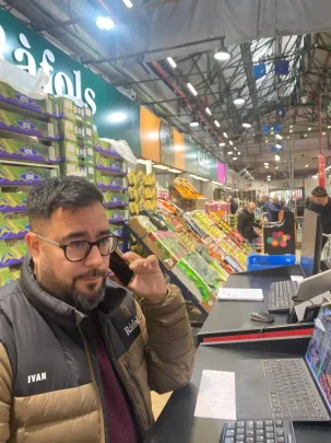 Iván Farré, Product Manager especializado en verduras, habla por teléfono en un puesto de mercado rodeado de cajas y expositores de frutas y verduras.