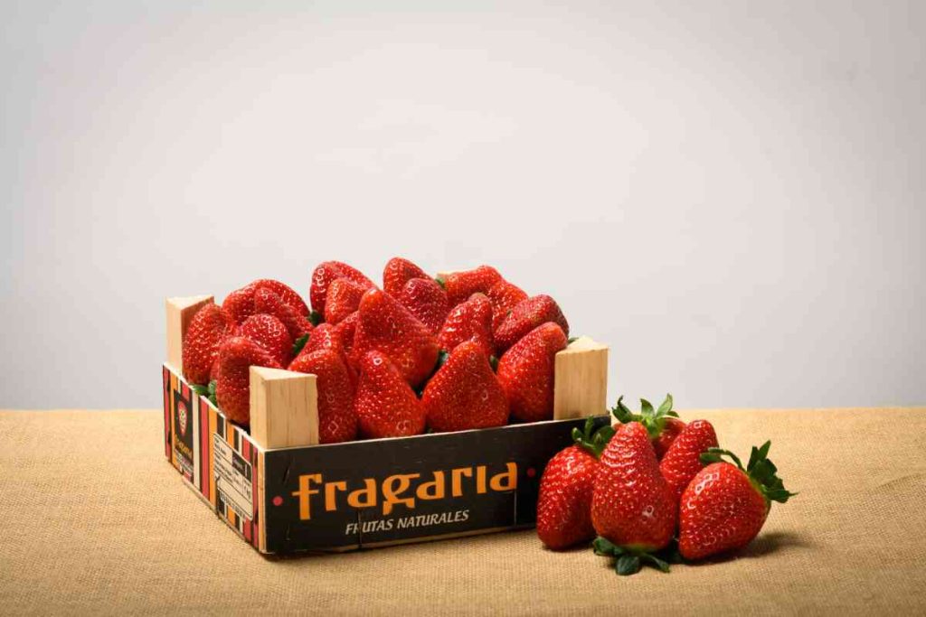 Caja de fresas con varias fresas delante sobre una superficie de tela. En la caja se lee "Fragaria Frutas Naturales".
